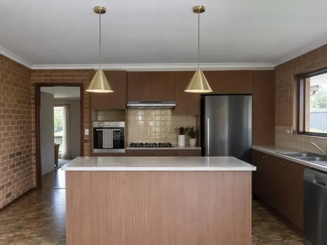 Kitchen with two brass pendant lights over an island, timber cabinetry and brick walls in Wyong